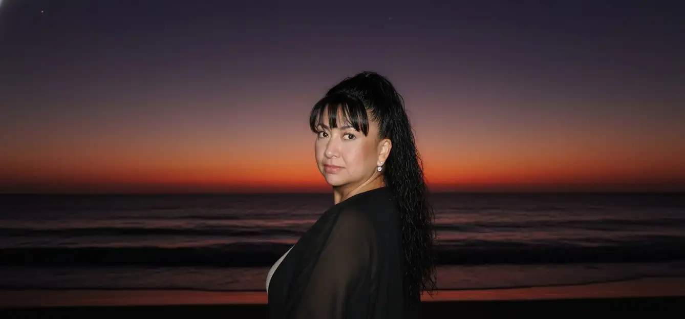 Hazme parecer que estoy en una playa oscura,

pero me sacaron una foto con flash y la cara brilla un poco. Al fondo, añade la puesta de sol, como si el sol se hubiera ocultado en un 98%, dejando un cielo rojo púrpura y casi oscuro. La imagen está tomada desde atrás y me han dicho que me gire para la foto. Llevo un traje negro playero. (Que la cara quede exactamente igual que en la foto).