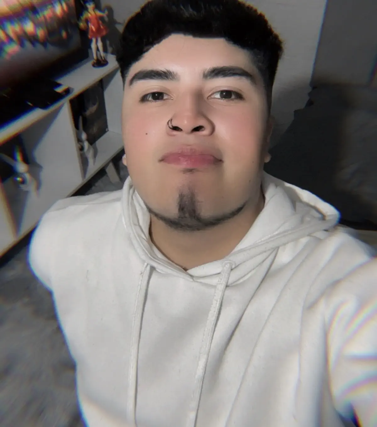 A high-contrast black and white photograph taken in a modern art gallery. The scene captures a candid, "stolen shot" moment of a young man like (my attached image) standing still in a moving crowd without changing his actual face. He is positioned slightly at an angle, not completely sideways with his face turned slightly, not completely facing the camera, but not completely sideways either. Framing from mid-thigh up (3/4 body shot) he is wearing a long dark coat with his hand in the pocket, standing out from the blurry crowd moving past him. The background wall is framed. Banksy's artwork is in a grid arrangement, slightly blurred for depth. The mood is moody, cinematic, and introspective.