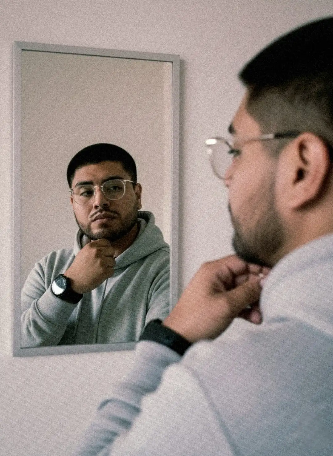 Make an image, keeping the person's facial features and face, with the following changes:
A realistic portrait photo inspired by a stylish male mirror selfie. The main focus is on the reflection in the mirror, while the real person in the foreground is softly blurred. The subject keeps his real facial and physical features, wearing glasses, a light gray hoodie (polera plomo claro) and a black wristwatch. He poses with one hand resting near his chin, gazing confidently at his reflection. Soft natural light from the side highlights the reflection’s face, creating a cinematic and modern aesthetic. The background is minimal, indoors, with neutral walls and a shallow depth of field. The overall tone is warm and slightly desaturated, shot with a portrait lens at f/2.8 using AE/AF lock, giving a professional vertical composition and ultra-realistic finish.